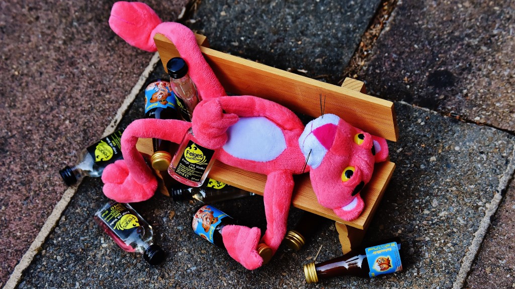 Vaaleanpunainen pantteri pehmolelu makaa puistonpenkillä tyhjiä pulloja ympärillään